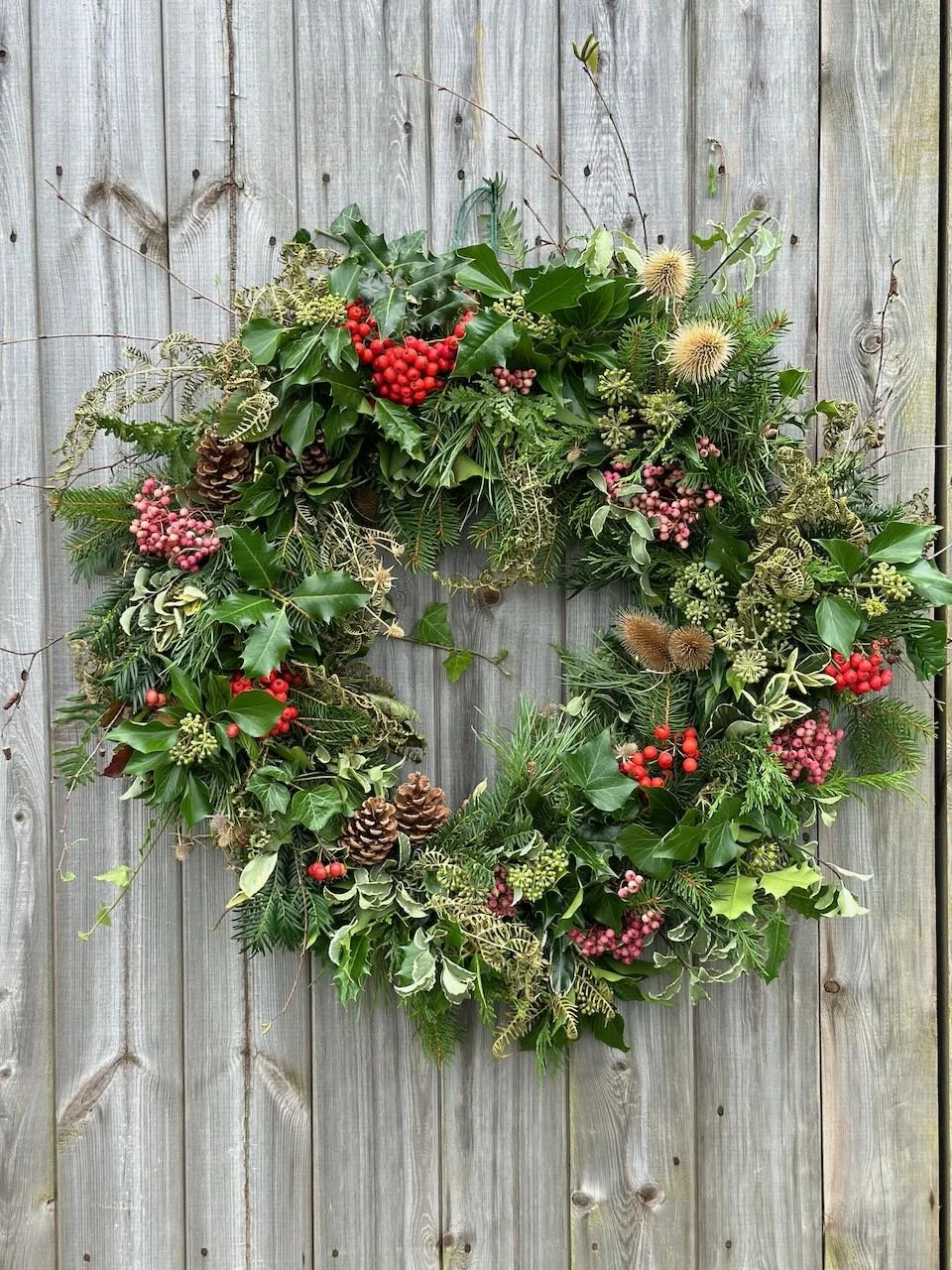 A festive Christmas wreath made of green leaves, pinecones, bright red berries, and other greenery hanging on a wooden fence.
