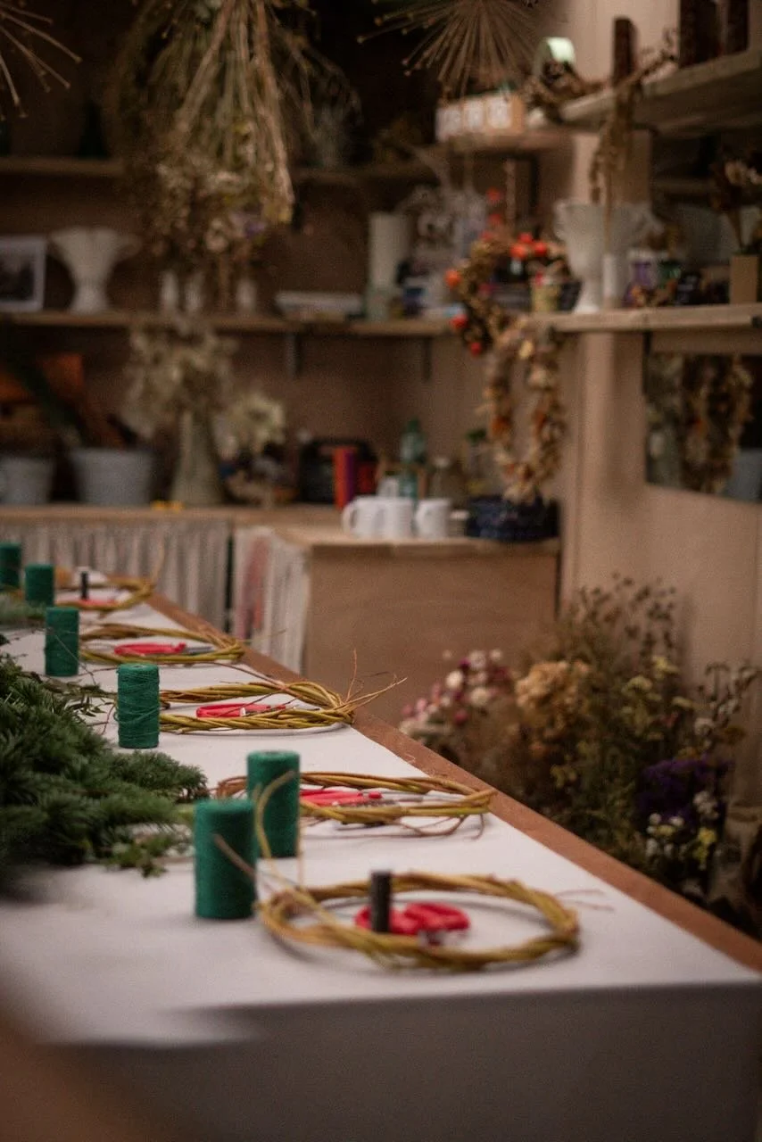 A wooden table with wreaths, red ribbons, and candles, set for a holiday craft or decoration project in a room filled with dried flowers and decorative items.