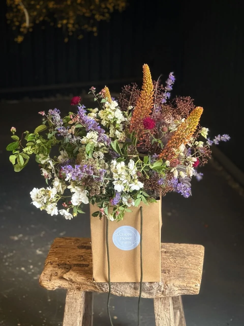 A bouquet of mixed flowers in a paper bag labeled 'Chippy Flower Farm' placed on a rustic wooden stool.