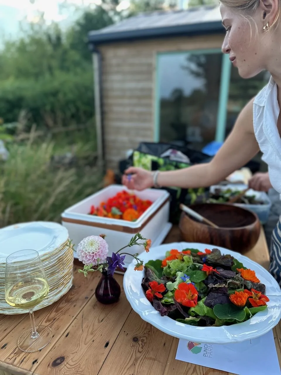 A woman is preparing a garden salad with mixed greens and edible flowers on a wooden table, with a glass of white wine, plates, a small vase with flowers, and a chest of vegetables in the background.