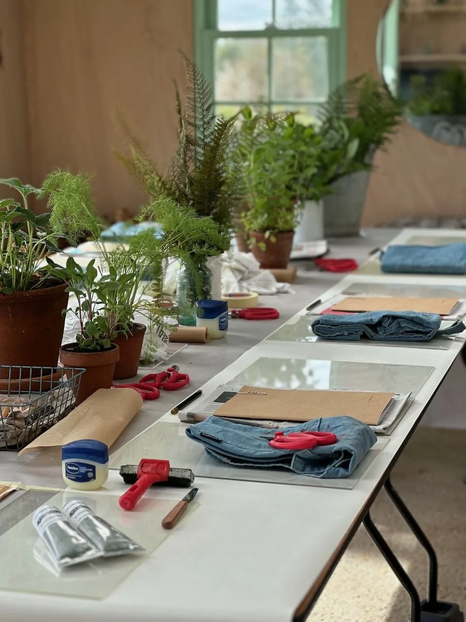 A workspace with potted plants, scissors, tape, markers, jeans, and other crafting materials on a white table, set near a window with trees outside.