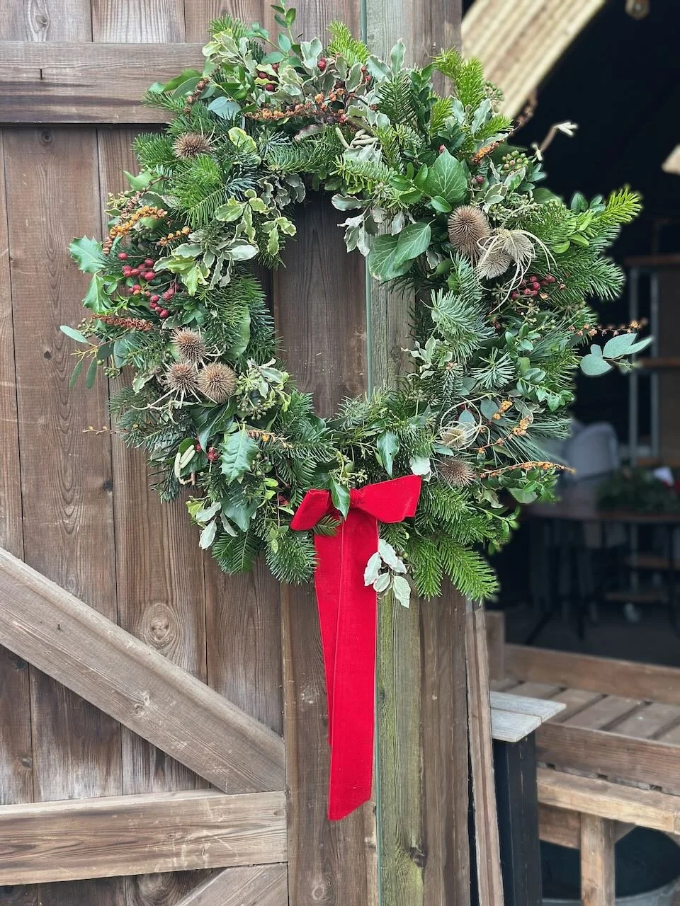 A Christmas wreath decorated with holly, pine, berries, and dried seed pods, with a red ribbon on a wooden door.