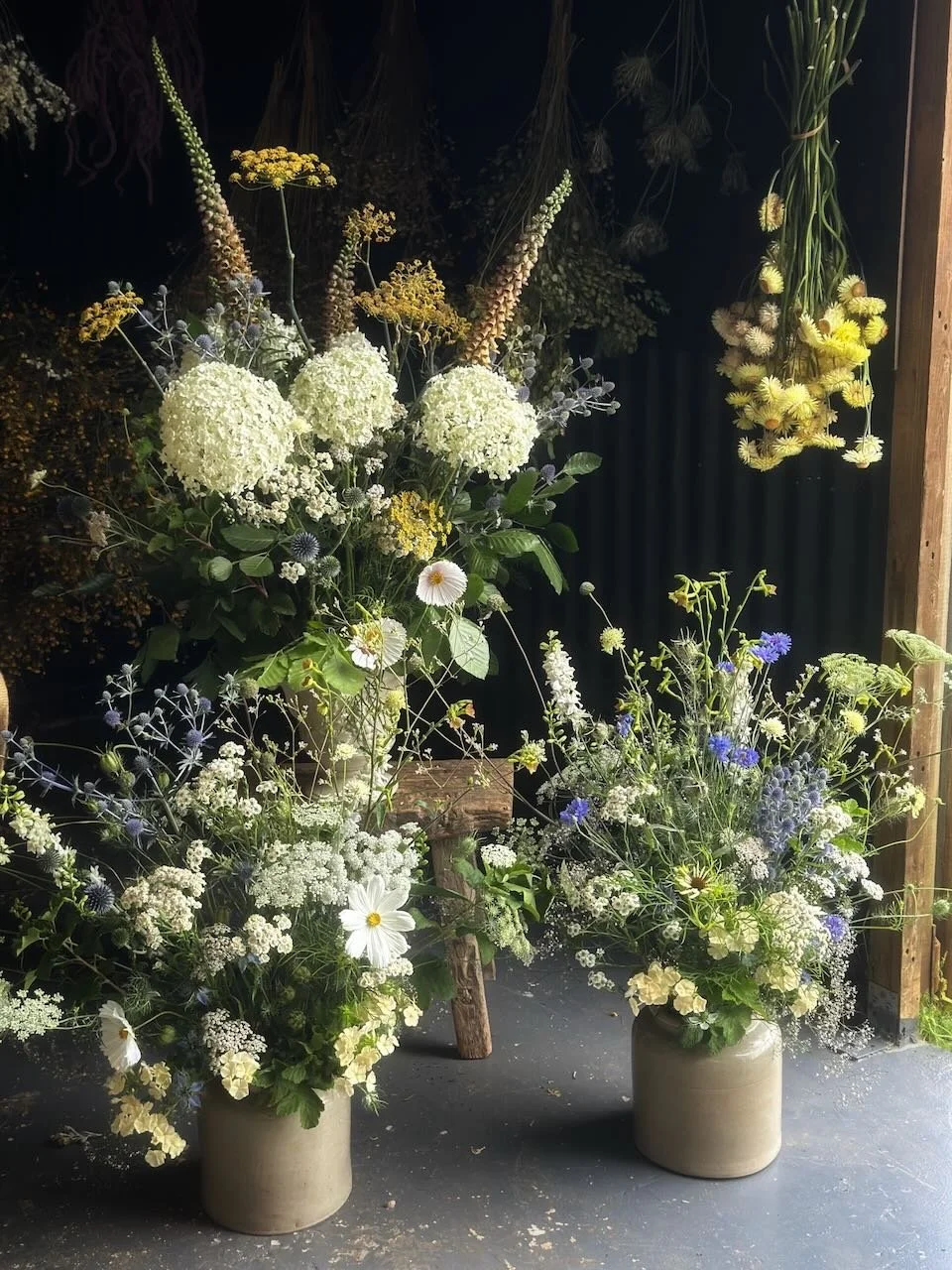 Two large flower arrangements in beige pots featuring white, yellow, purple, and blue flowers placed on a dark floor with a black background.
