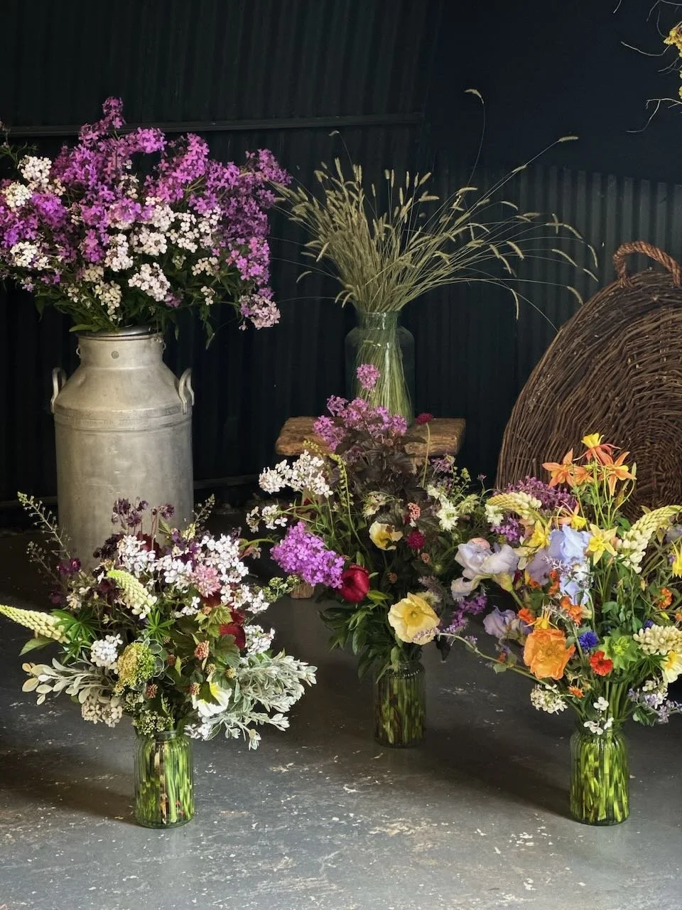 Various colorful flower bouquets in green glass vases in front of a black background with a wicker basket and additional vases of plants.