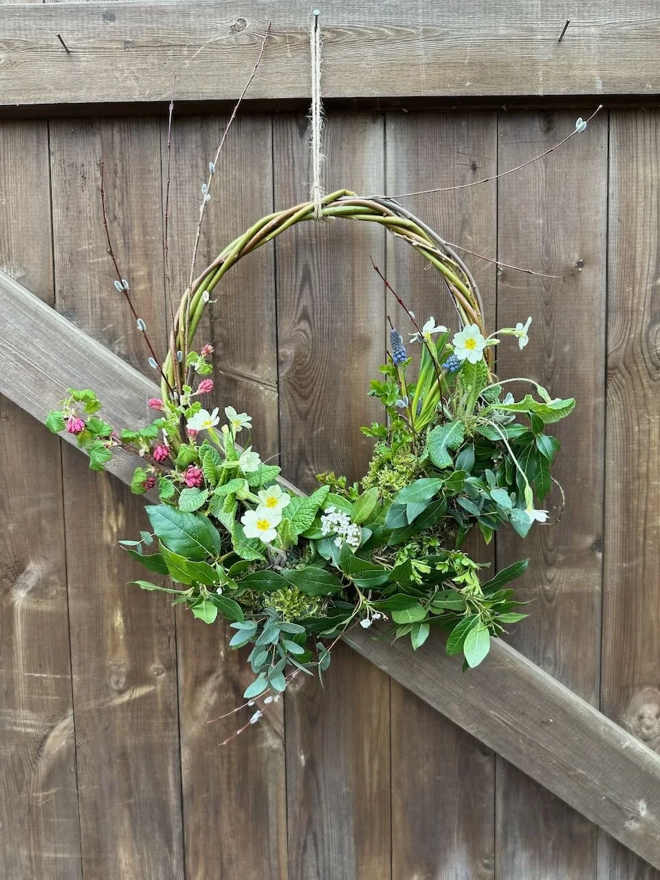 A decorative wreath made of intertwined branches and various green leaves, adorned with small white, pink, and blue flowers, hanging on a wooden fence.