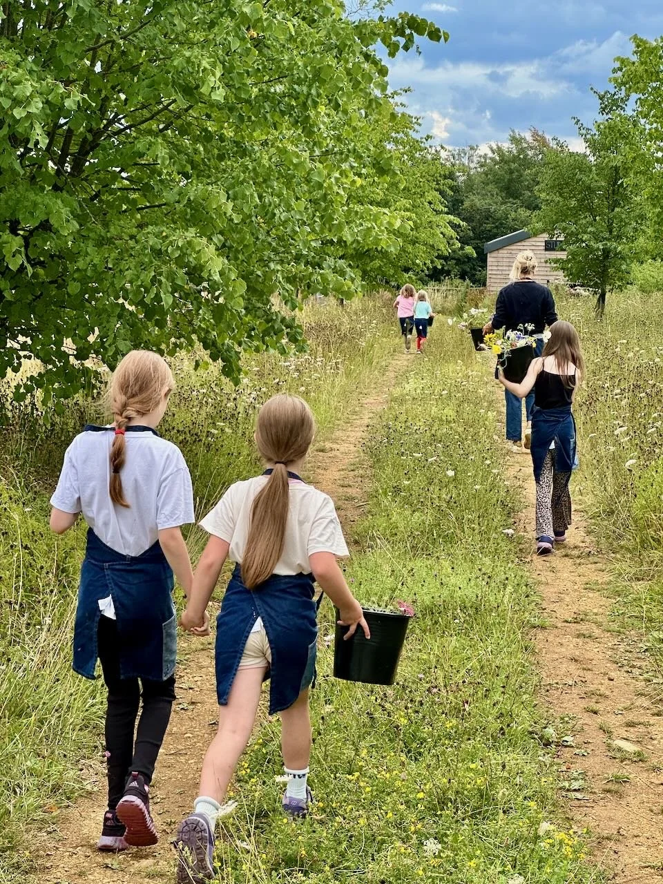 Children and adults walking along a dirt path through a grassy, overgrown field, with trees on the sides and a small building in the distance, during daytime.