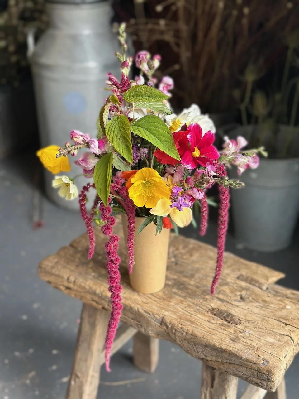 Colorful flower arrangement in a beige vase on a rustic wooden stool, with a background including a metal watering can and garden pots.