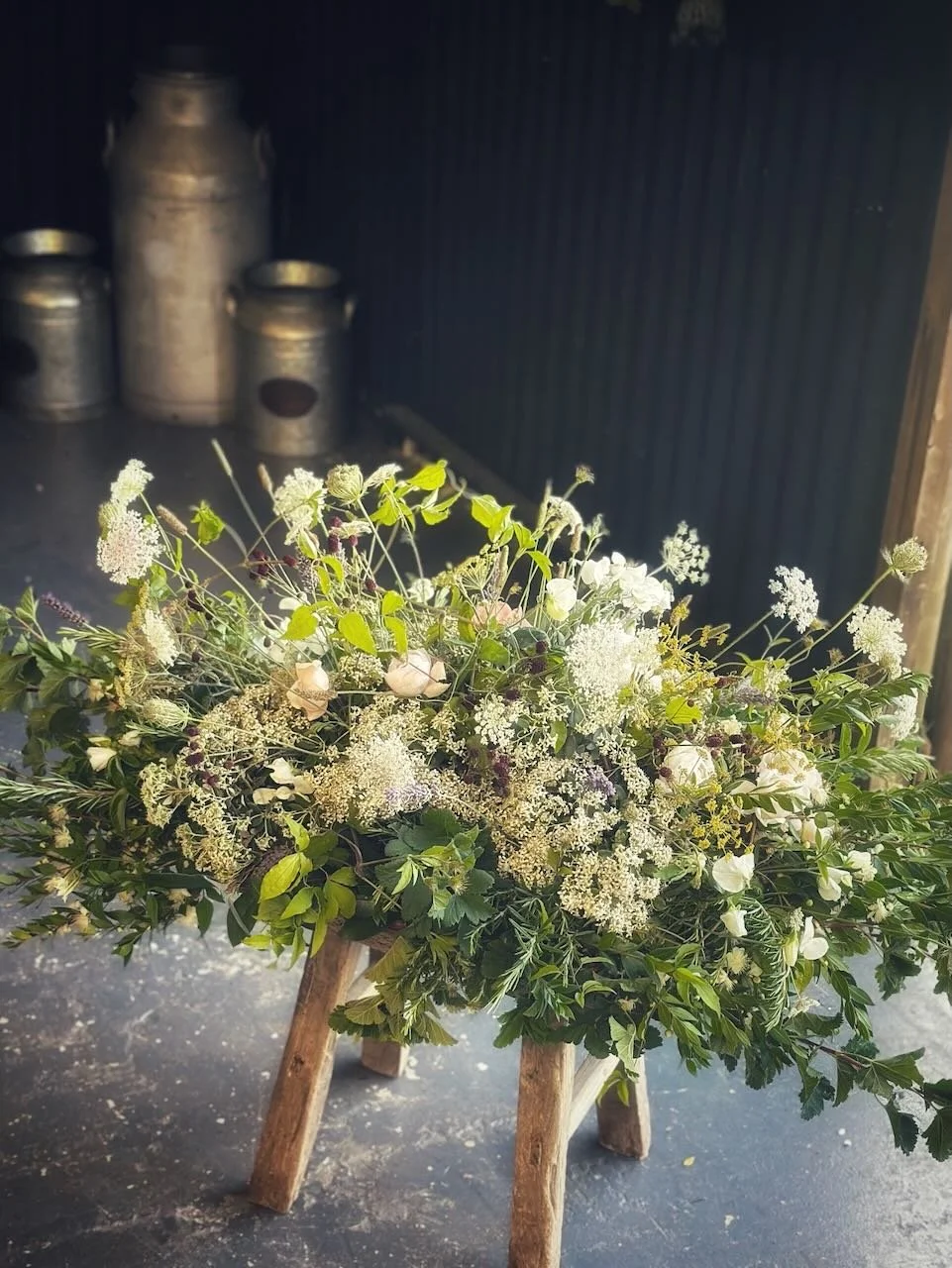A floral arrangement with white flowers and greenery on a small wooden stand, set against a black wall with metal milk cans in the background.