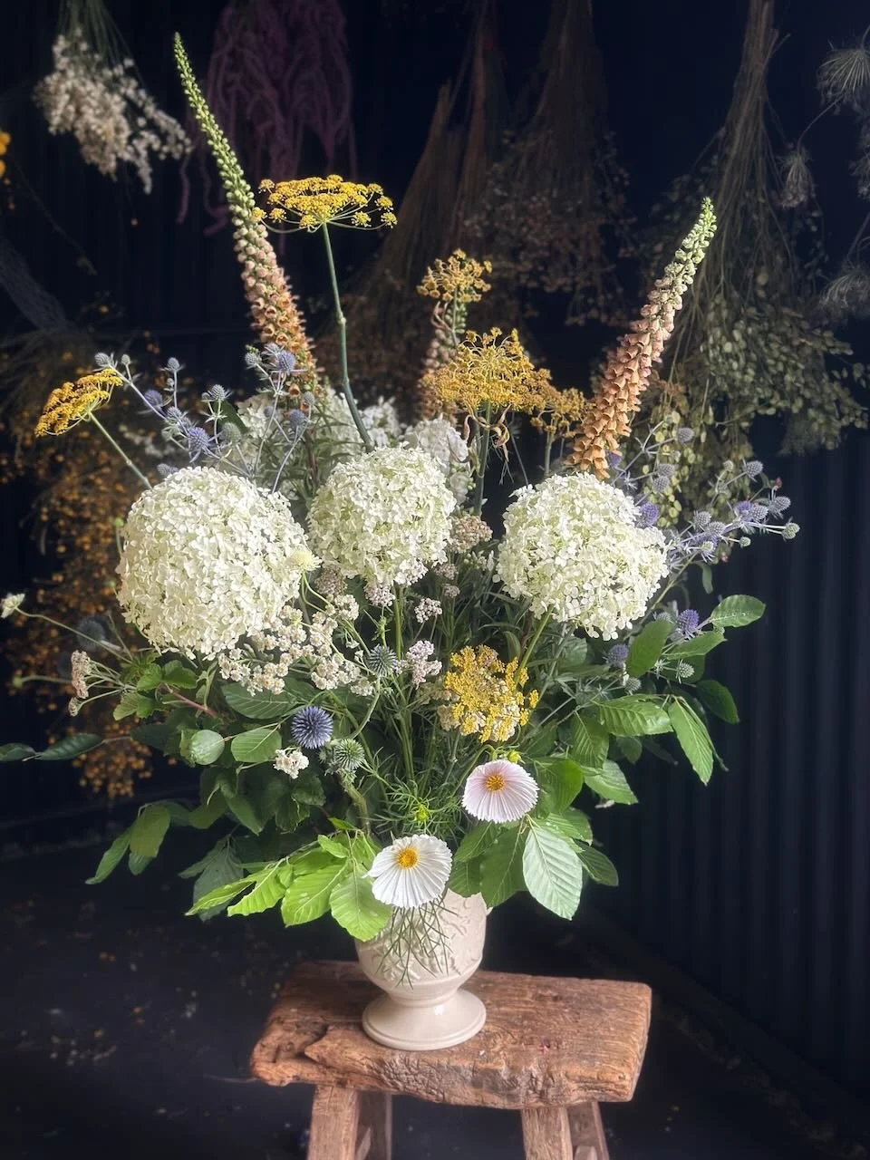 A floral arrangement with white hydrangeas, daisies, and various wildflowers in a cream-colored vase on a wooden stool.