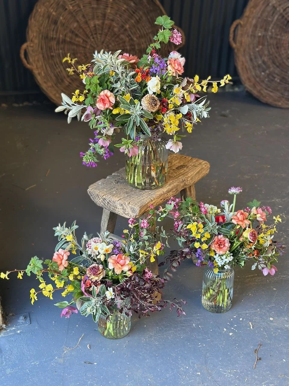 Three glass vases filled with colorful mixed flowers, placed on a rustic wooden stool and the floor, with woven baskets in the background.