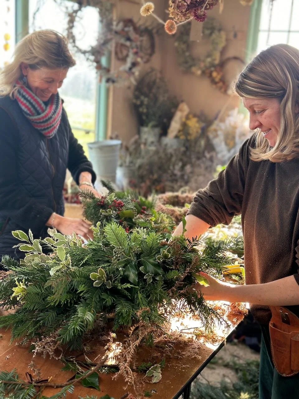 Two women crafting a holiday floral arrangement with evergreen branches, holly berries, and other winter foliage indoors near a window.