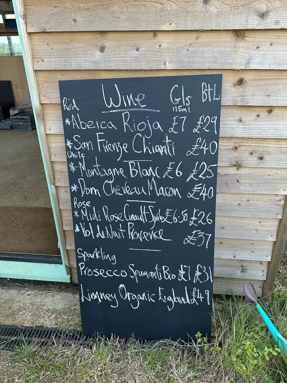 Chalkboard sign listing wines and prices outside a building, with a wooden exterior, and some grass and a garden tool at the bottom right.