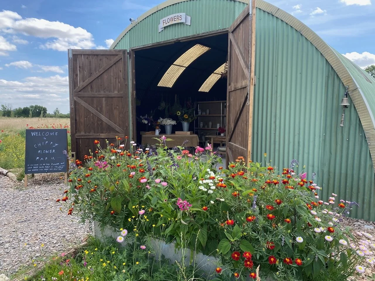A green curved metal barn with open double doors revealing a flower shop inside. Outside, colorful flowers grow in a large container, and a black chalkboard sign welcomes visitors to the flower farm. The sky is partly cloudy with a rural landscape in