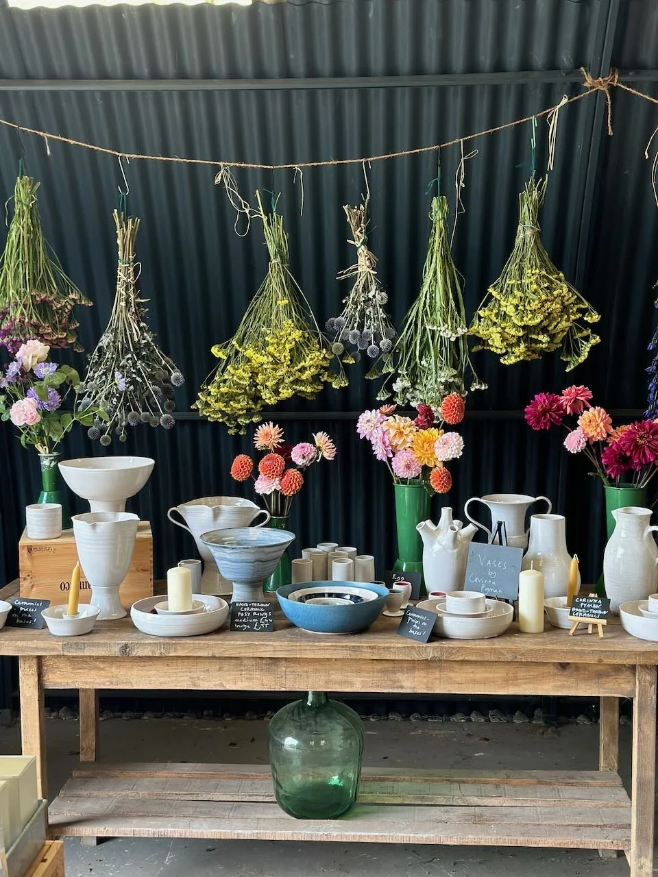 Display of various ceramic vases and flower arrangements on a wooden table in a shop, with dried flower bundles hanging above against a dark curtain background.