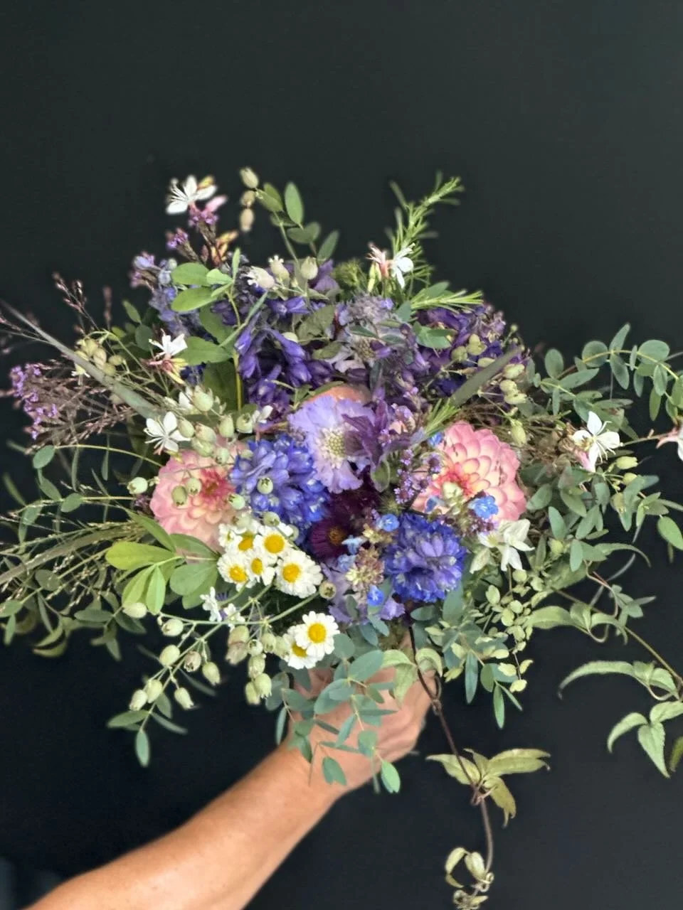 Hand holding a colorful bouquet of mixed wildflowers against a dark background.