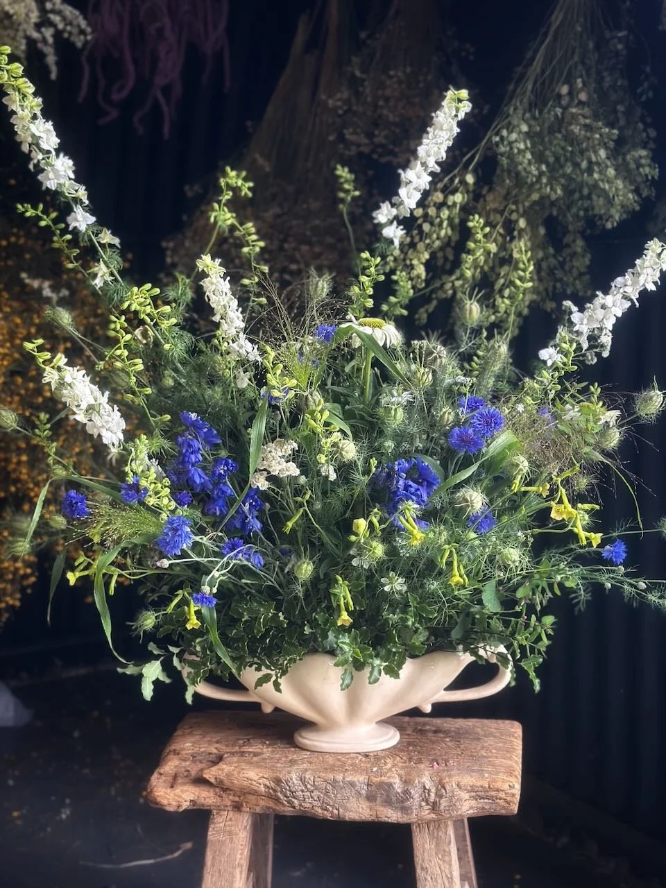 A large white ceramic vase filled with an assortment of white and blue flowers and greenery, placed on a rustic wooden stool against a dark background.