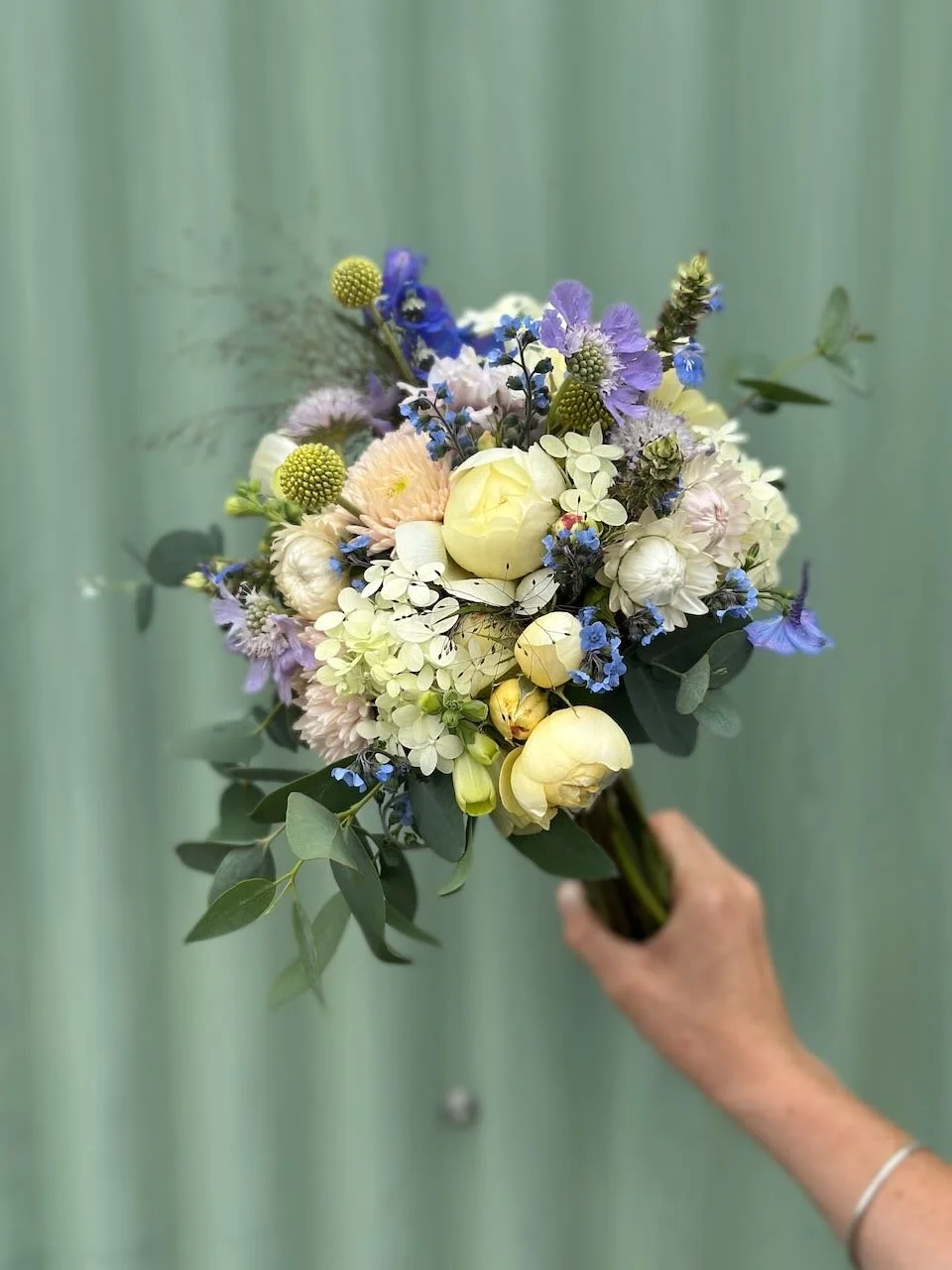 A person holding a bouquet of mixed flowers and greenery against a blurred green background.