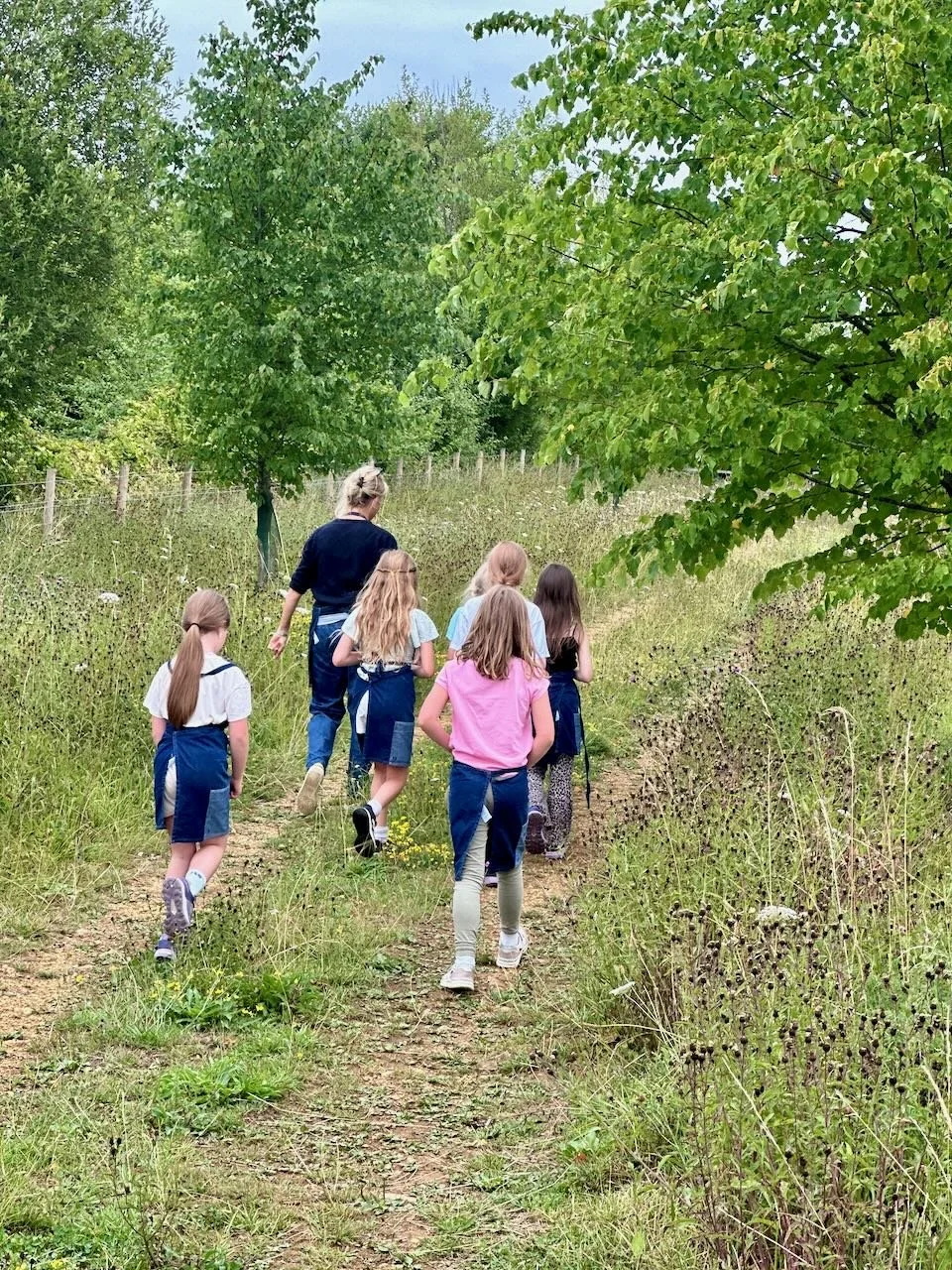 A group of children walking along a dirt path in a lush green outdoor setting, accompanied by an adult.