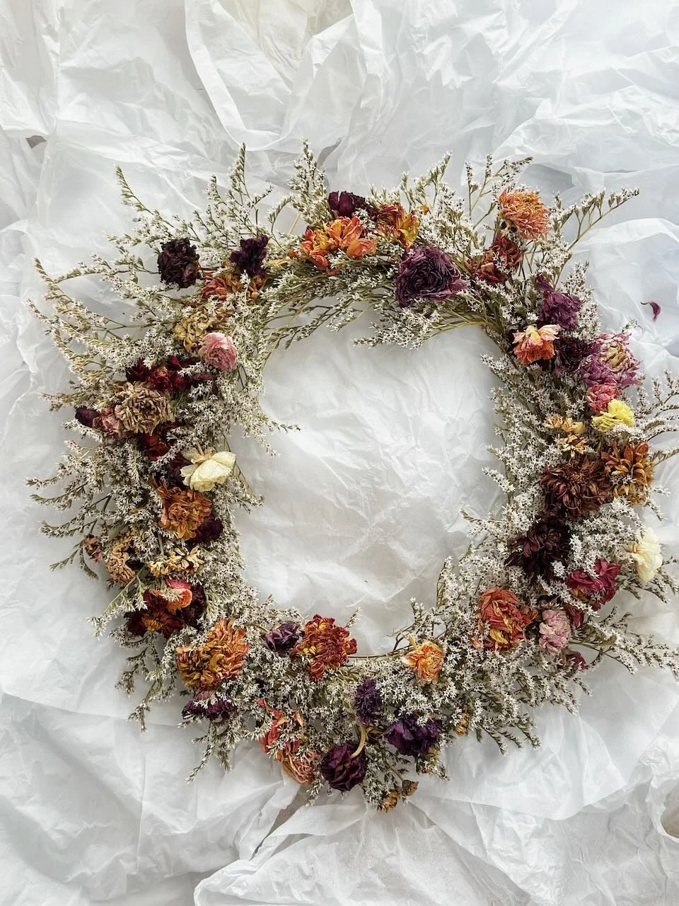 A circular floral wreath made of dried flowers in shades of purple, orange, yellow, and white, resting on crumpled white tissue paper.