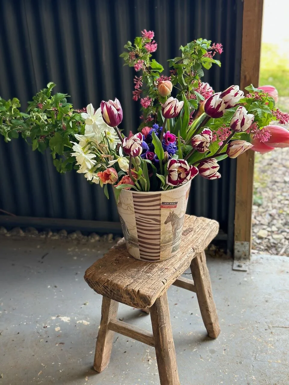 A bouquet of pink, white, and purple tulips and other flowers in a decorated paper bucket on a rustic wooden stool, set against a dark and outdoor background.