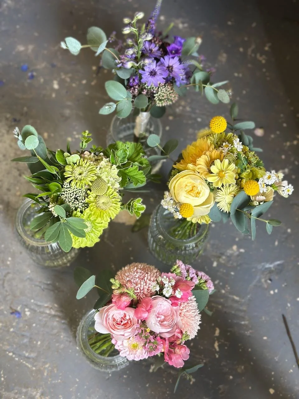 Four small glass vases containing various colorful flower bouquets placed on a concrete surface. The bouquets include purple, yellow, pink, and mixed flowers with green foliage.