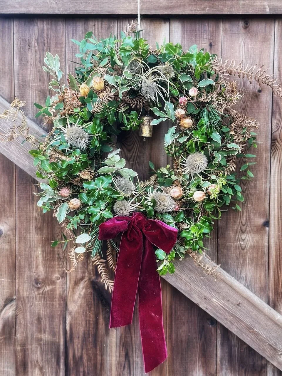 A holiday wreath made of green leaves, dried flowers, and seed pods, decorated with a red velvet bow, hanging on a wooden fence with diagonal support.