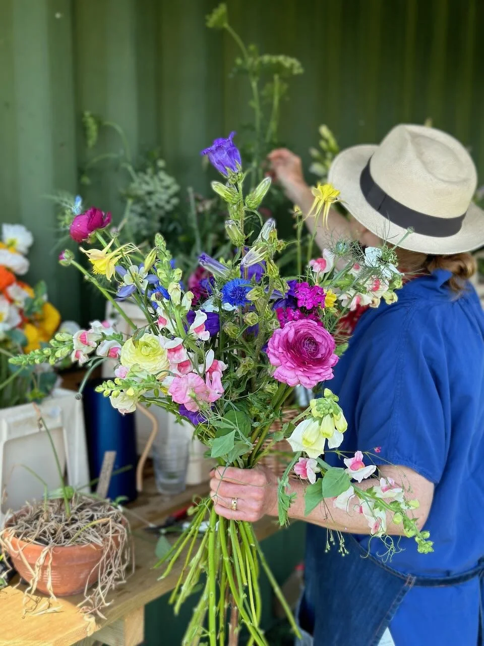 A person in a blue shirt and wide-brimmed straw hat holding a colorful bouquet of flowers, with a floral shop setting in the background.