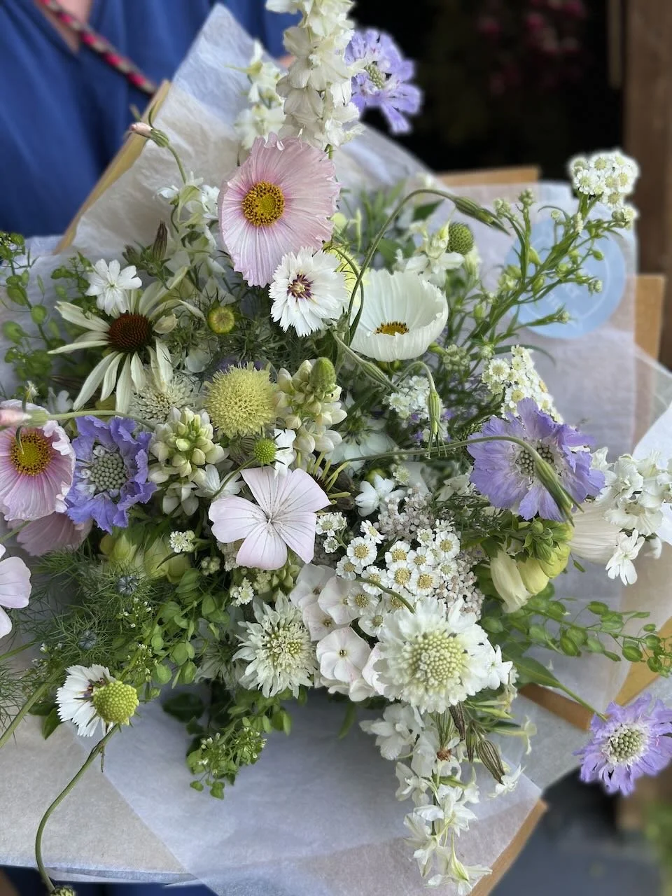 A bouquet of mixed flowers including white, pink, and purple blooms with various green foliage, wrapped in paper.