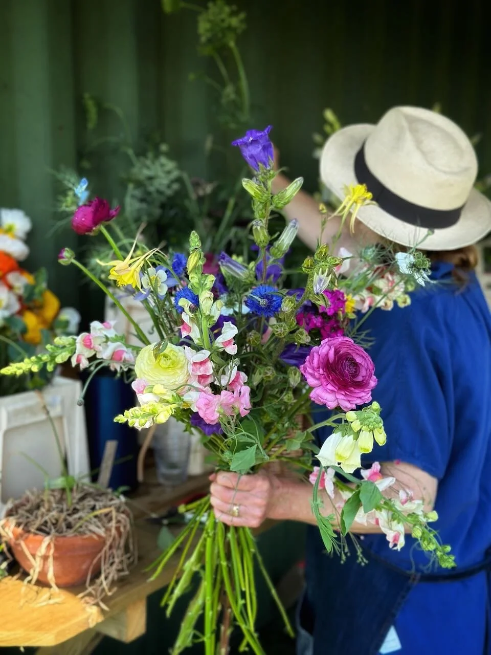 Person wearing light-colored hat and blue shirt holding a colorful bouquet of fresh flowers, with a green wall in the background and gardening supplies on a wooden table.