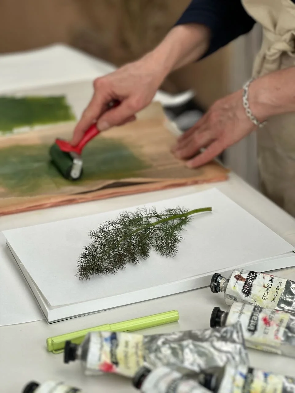 A person painting on paper with a roller brush, with a green leaf placed on top of a white sheet of paper on a table. Several tubes of ink and a green pen are also on the table.