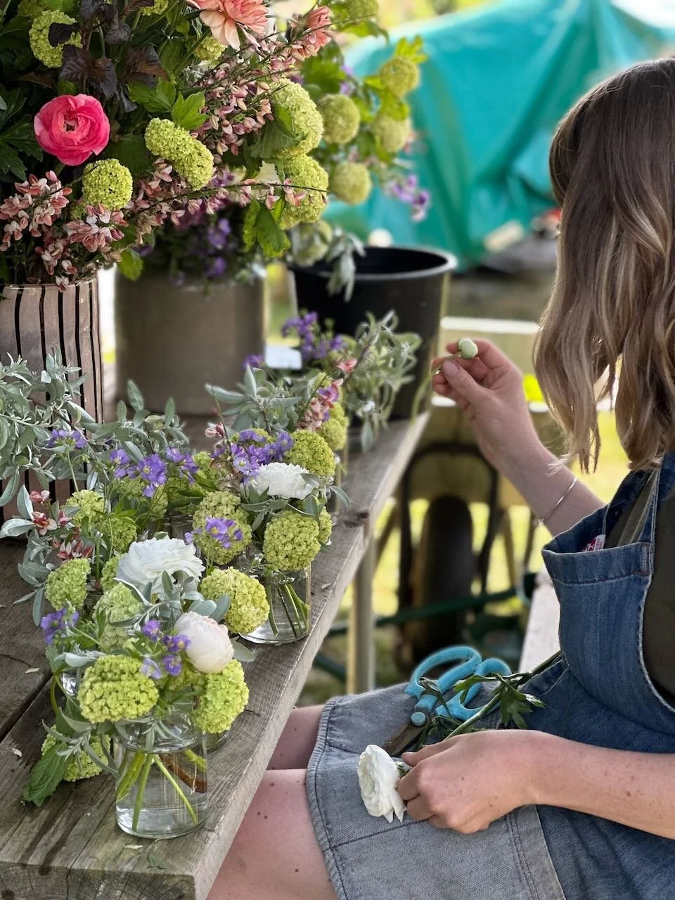 Woman arranging colorful flowers in small glass vases on a wooden table at a flower arranging event.