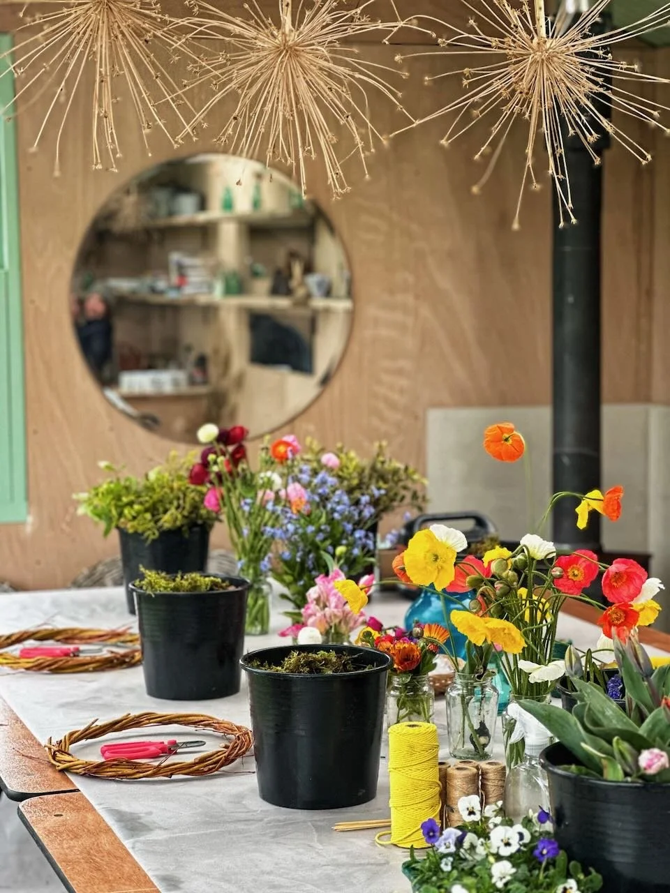 Table with various flower arrangements, scissors, and wreaths in a workshop or floral studio setting.