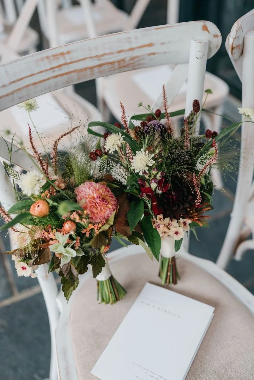 Two bouquets of mixed flowers resting on a white wooden chair with a pinkish cushion, next to a white card on the seat.