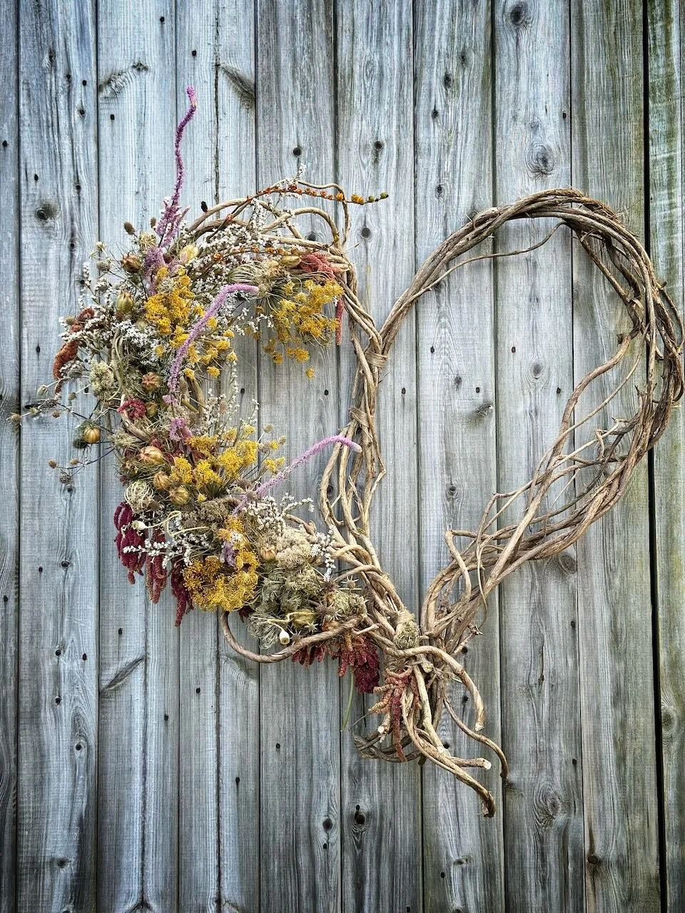 Heart-shaped wreath made of intertwined natural twigs decorated with dried flowers, hanging on a weathered wooden fence.