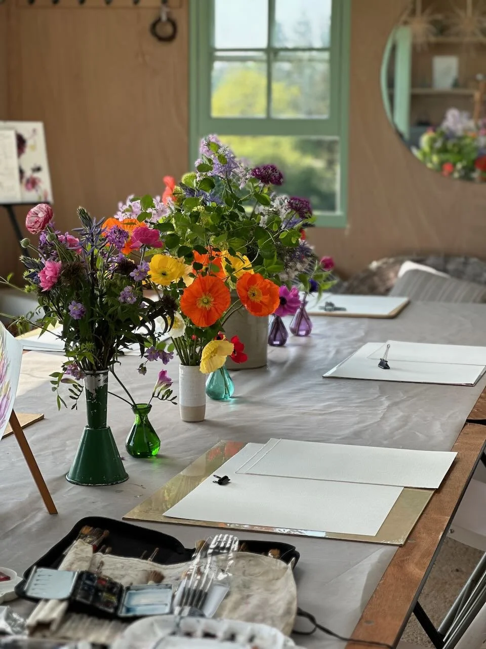 Table with colorful flowers in vases and art supplies, set in a cozy room with a window and wall mirror.