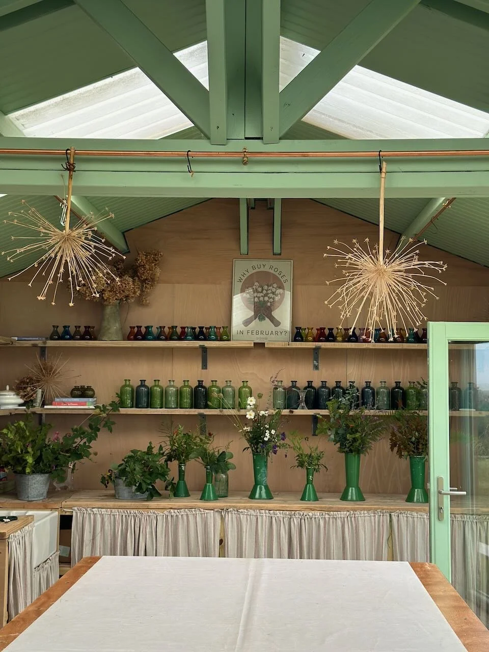 Interior of a flower shop or boutique with green vases and plants on wooden shelves, hanging decorative seed pods, a framed poster, and a door to the right.