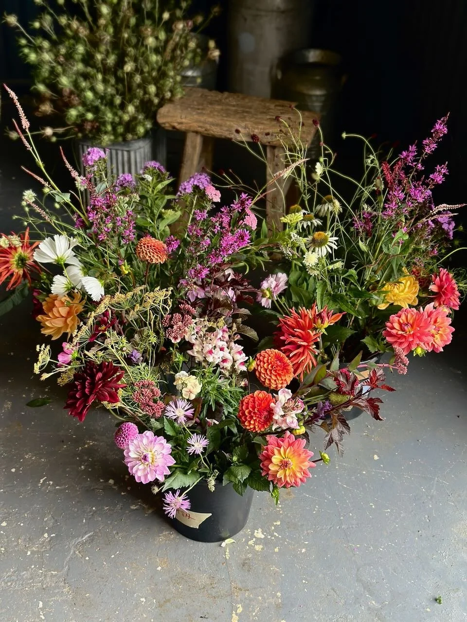 A colorful bouquet of various flowers, including dahlias, daisies, and other vibrant blossoms, arranged in a black pot on a gray surface.