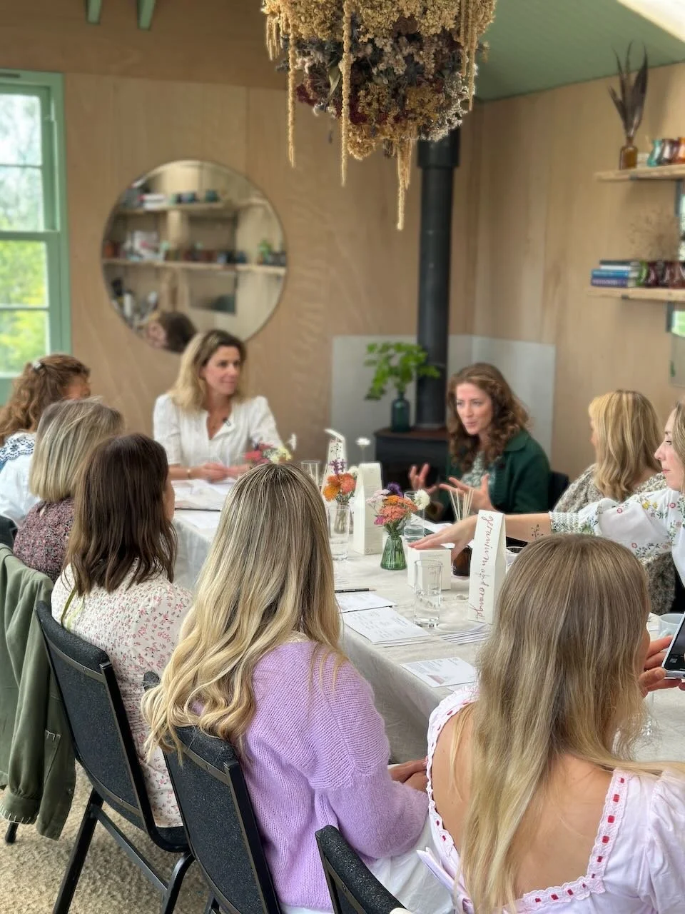 Women gathered around a dining table engaged in conversation, with a woman speaking, in a cozy room with windows, bookshelves, and dried flower decor hanging from the ceiling.