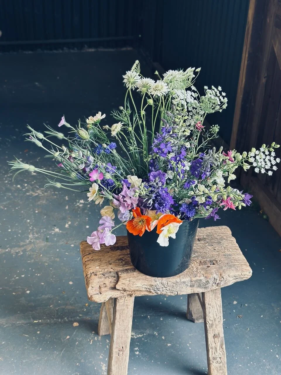 A black pot filled with various colorful flowers, placed on a small wooden stool outdoors.