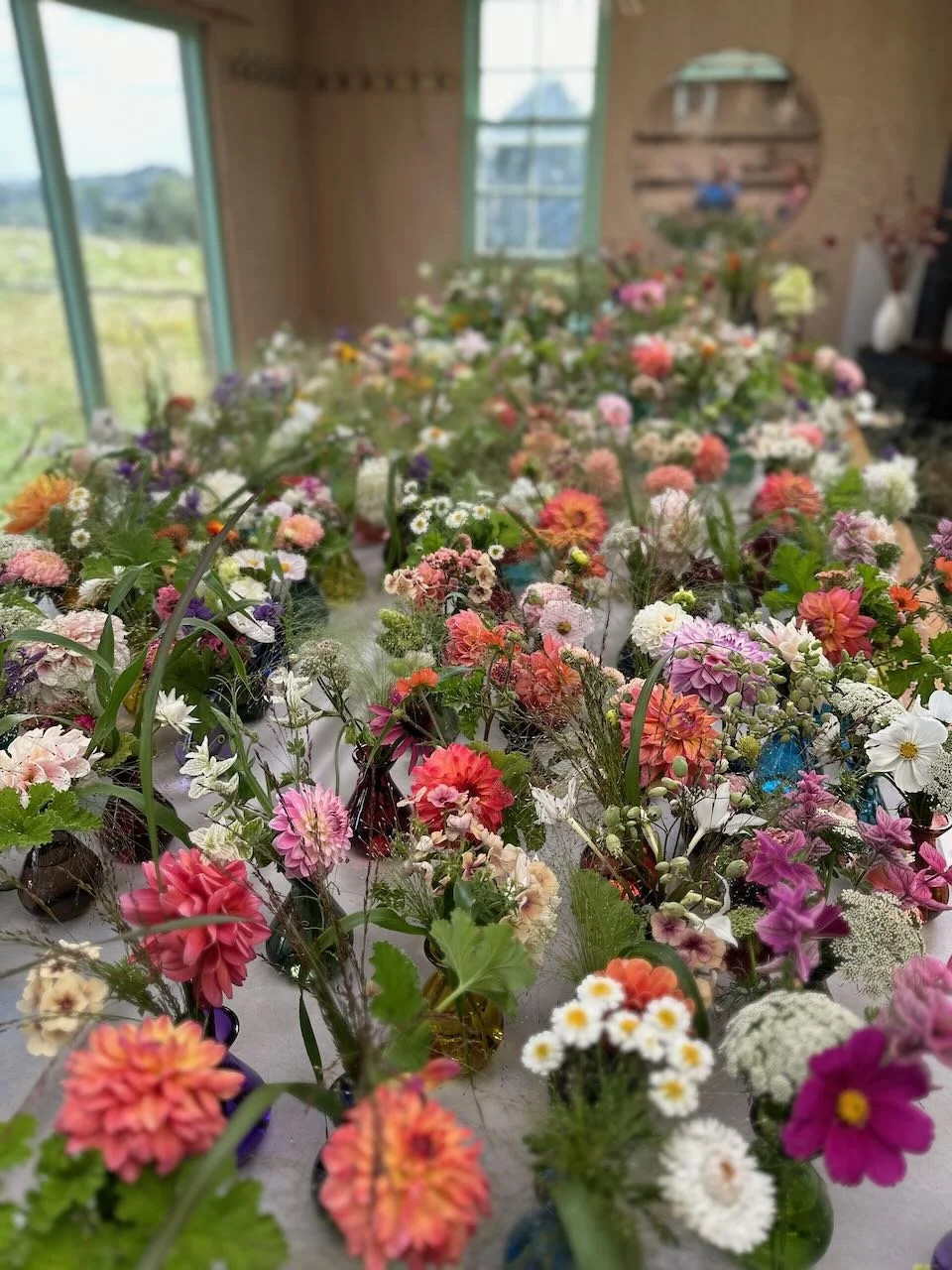 A room filled with numerous colorful flowers arranged in small vases on a long table, with windows and a mirror reflecting the interior.