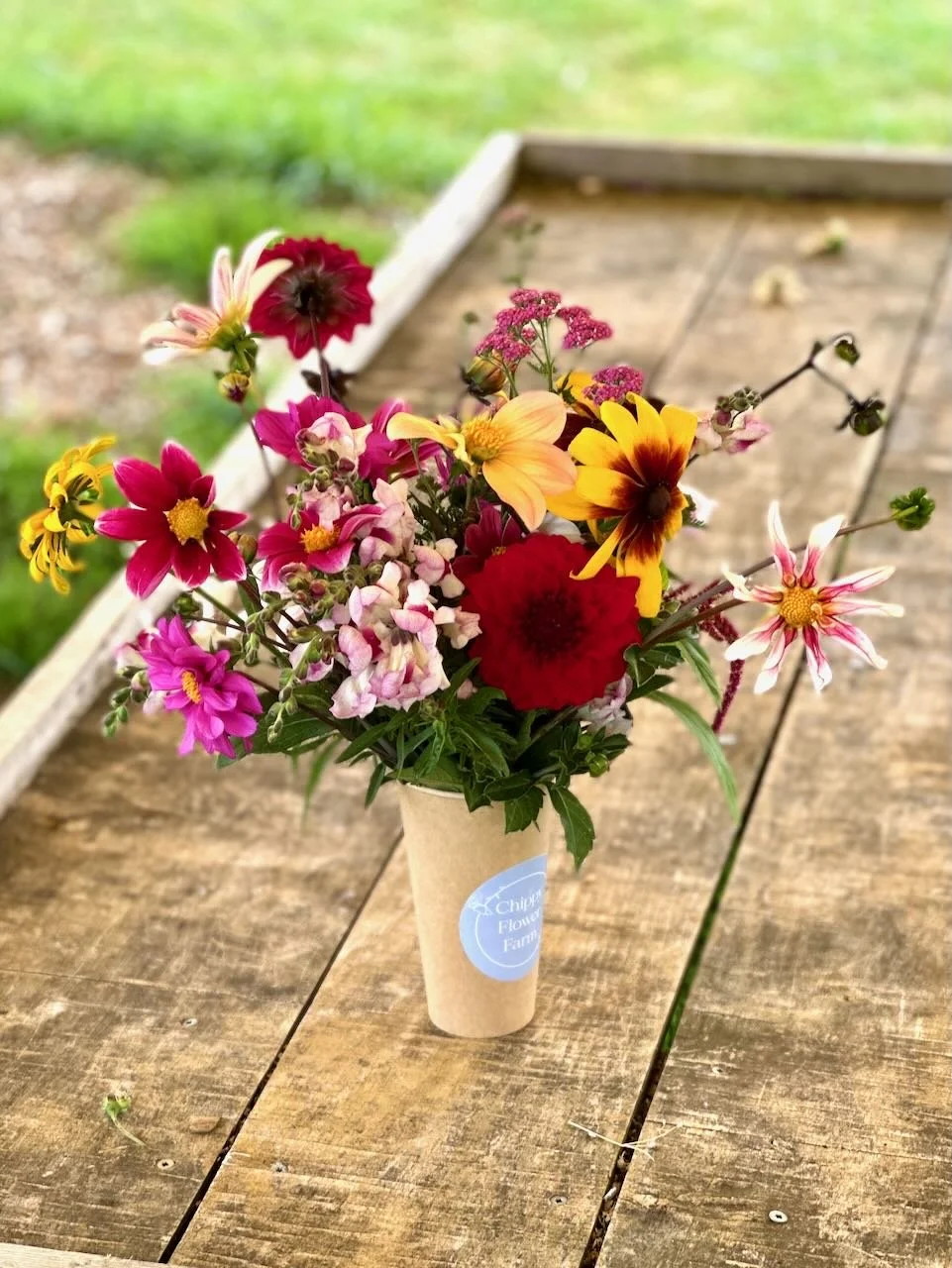 A small paper cup filled with colorful mixed flowers, placed on a wooden outdoor table.