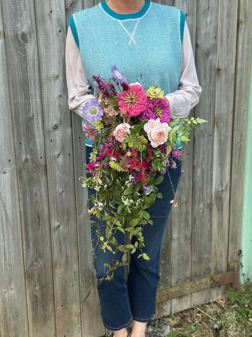 A person standing in front of a wooden fence, holding a large bouquet of colorful flowers with pink, purple, yellow, and white blossoms and greenery, wearing a blue sleeveless sweater over a white long-sleeve shirt and jeans.