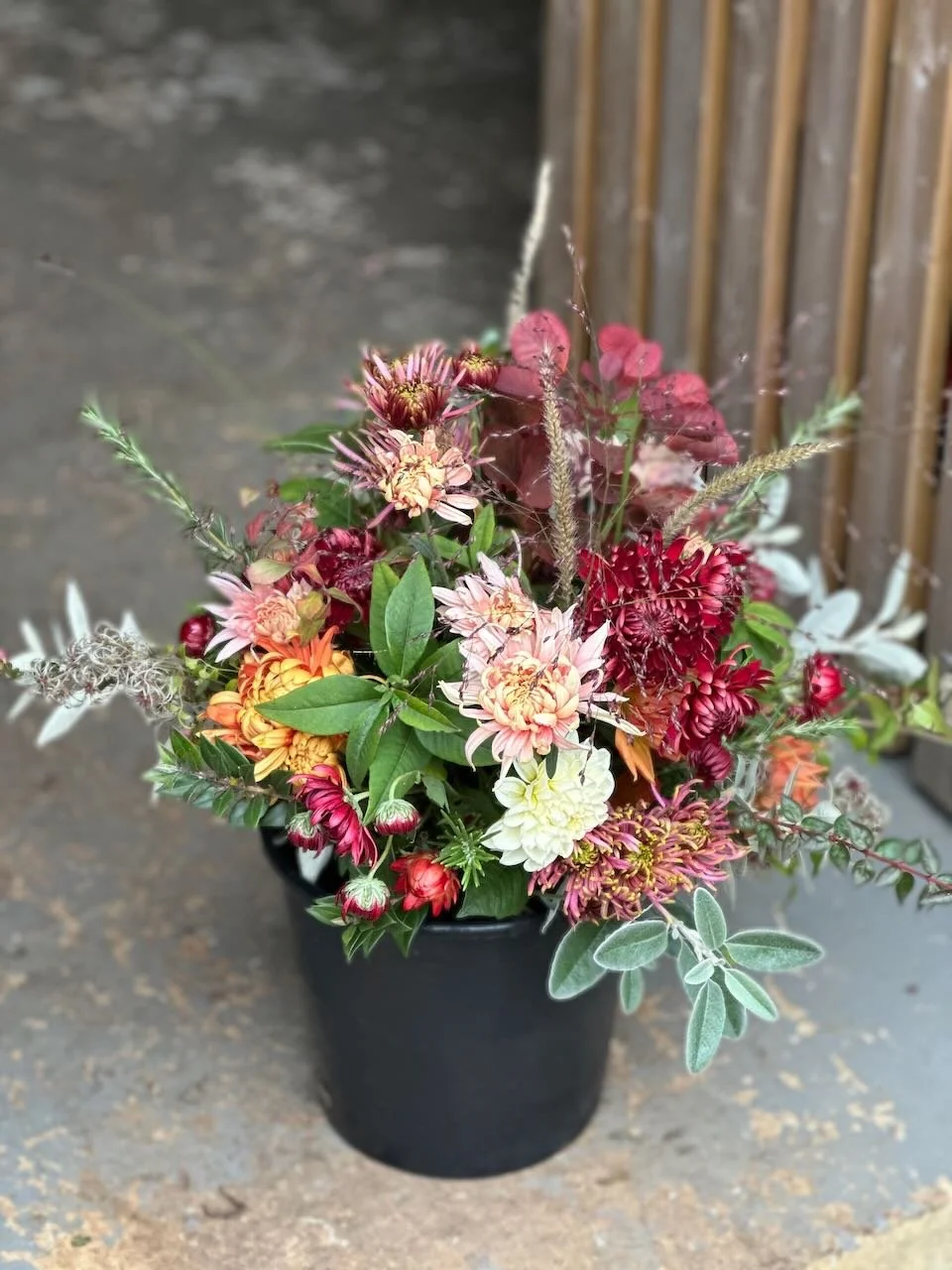 A black pot filled with various colorful flowers and greenery, set on a concrete surface with a wooden fence in the background.