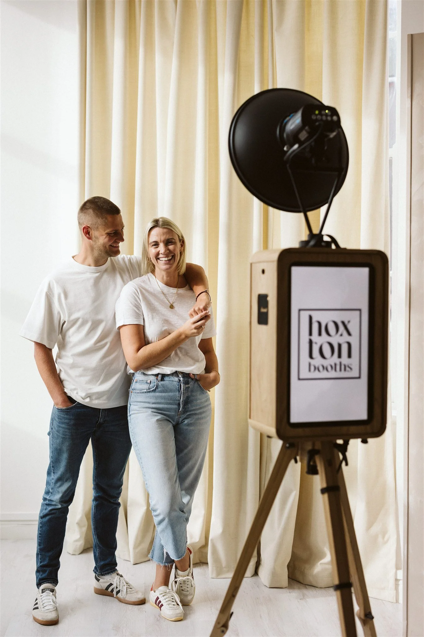 A young man and woman stand together in a photo booth, smiling and laughing. The woman has her arm around the man's shoulder, and they appear to be enjoying themselves. The booth has a sign that says 'hox ton booths' and is set against beige curtains.
