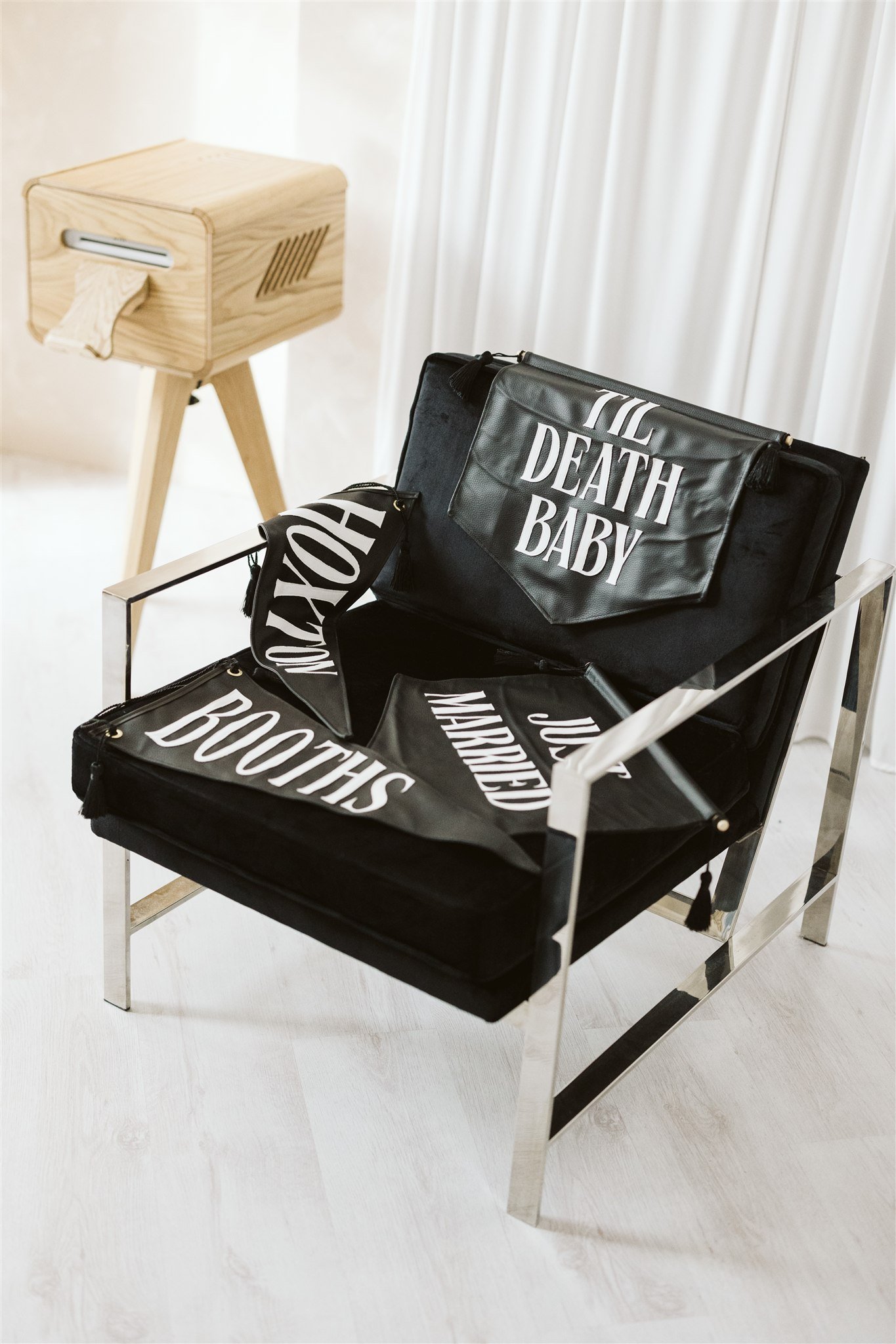 Black tote bags with white text reading 'TIL DEATH BABY' and 'BOOTHS' on a modern metal chair, and a wooden photo booth print station in the background.