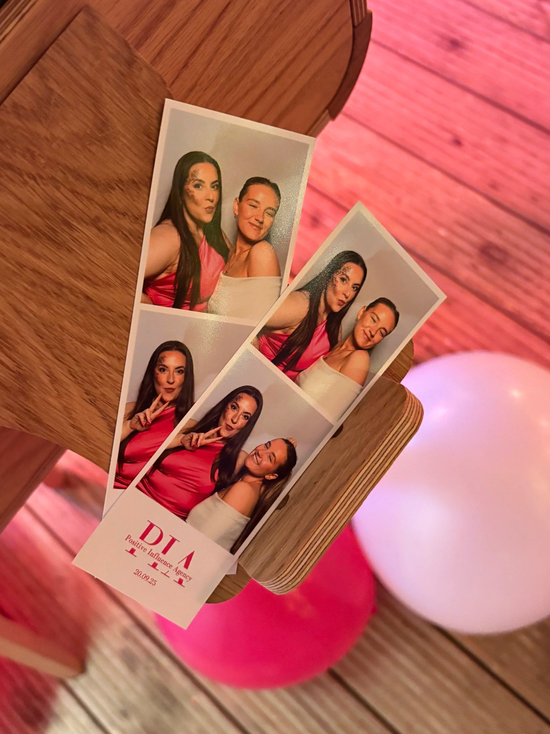 Photo booth strip with four pictures of two women, one with long dark hair and the other with shorter, lighter hair, posing together in a pink top. The strip has the words 'PIA Positive Influence Agency 20.09.25' at the bottom.