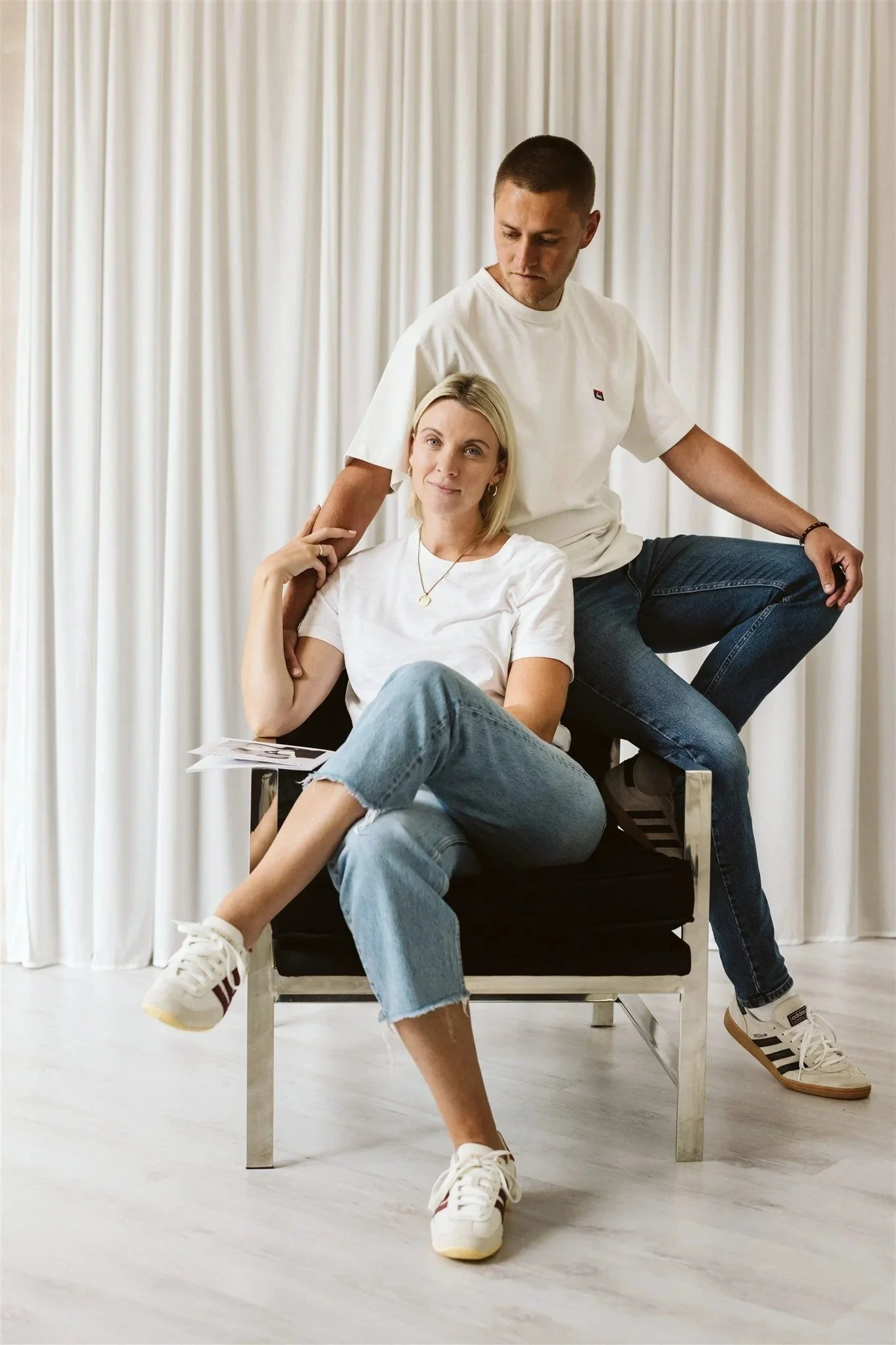 A woman sitting on a black chair with a man behind her, both dressed casually, in front of a white curtain, with the woman holding a piece of paper.
