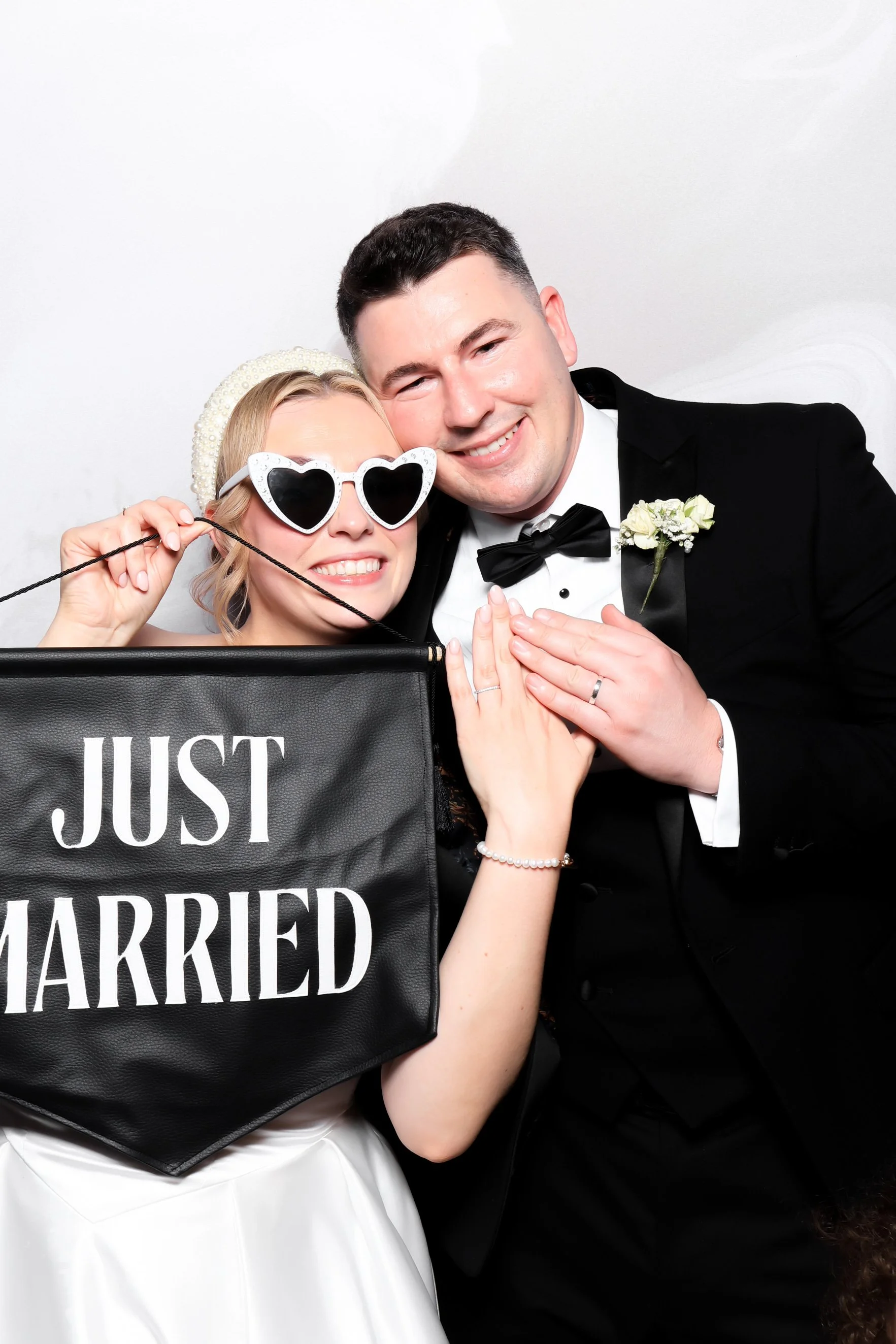 A newlywed couple dressed in wedding attire, smiling and holding a 'Just Married' sign, posing for a photo with some fun accessories.