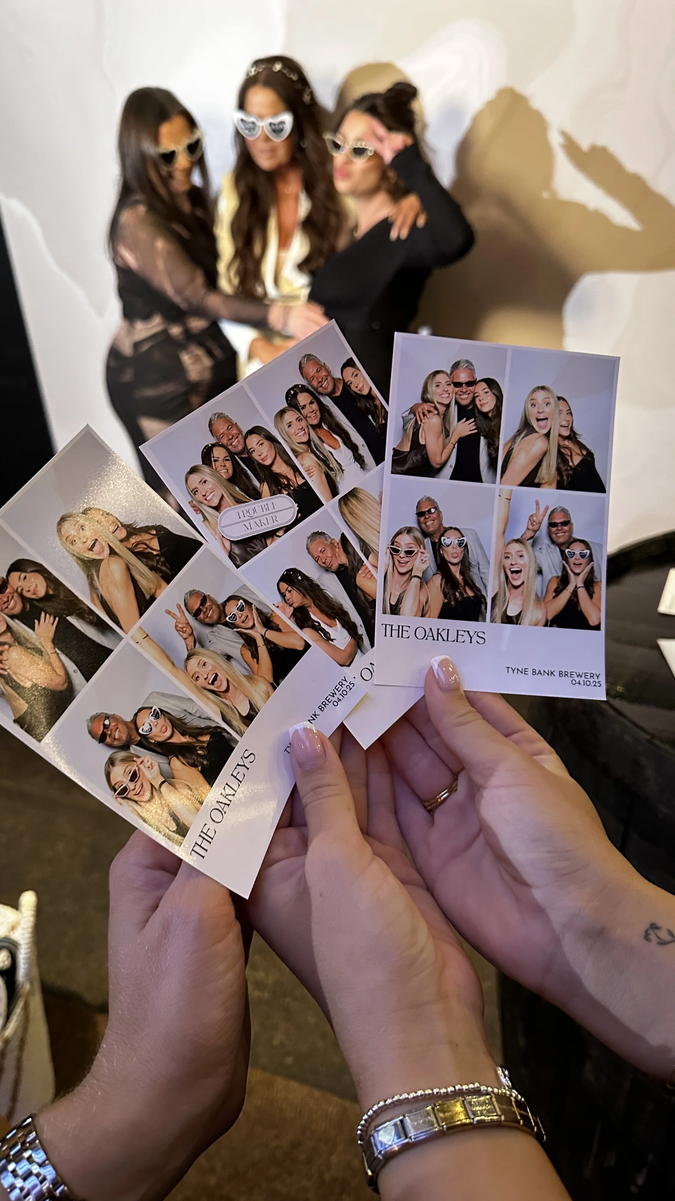 A person holding photo booth pictures of a group of people at an event, with a larger group photo in the background of three women posing together, all wearing sunglasses.