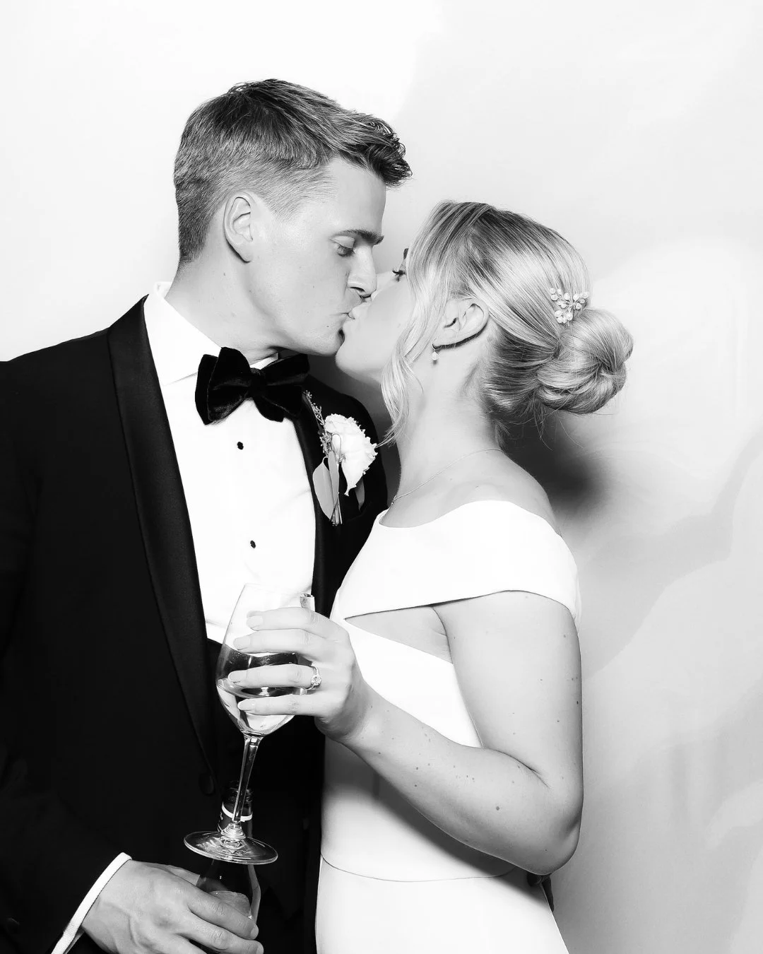 Black and white photo of a newlywed couple kissing, the groom in a tuxedo with a bow tie and boutonnière, holding a glass of champagne, and the bride in a wedding dress with hair in an updo decorated with flowers.